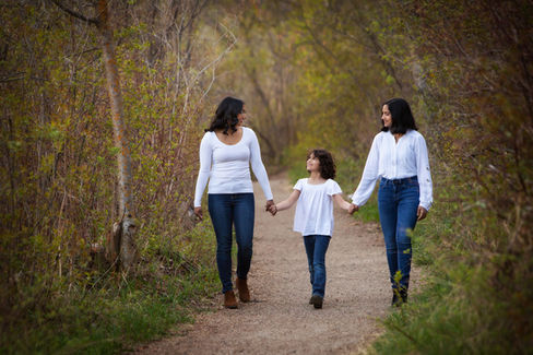 Two women and a child, all in white tops and jeans, walk hand-in-hand on a forest path. They are smiling, surrounded by greenery. Viewpoint Photography Edmonton