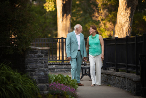 Elderly couple walking hand in hand, smiling, in a lush park. He wears a light blue suit, she wears a teal top. Peaceful, sunny setting. Viewpoint Photography Edmonton