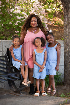 A woman in a pink dress stands with three smiling girls in blue dresses by a tree and bench, with blooming flowers in the background. Viewpoint Photography Edmonton