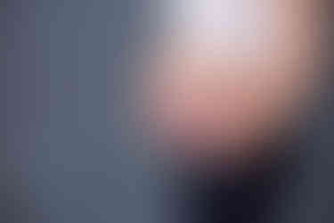 Pregnant woman in jeans holds pink "Ava" sign over belly. Wearing a white top, against a textured blue-grey background.