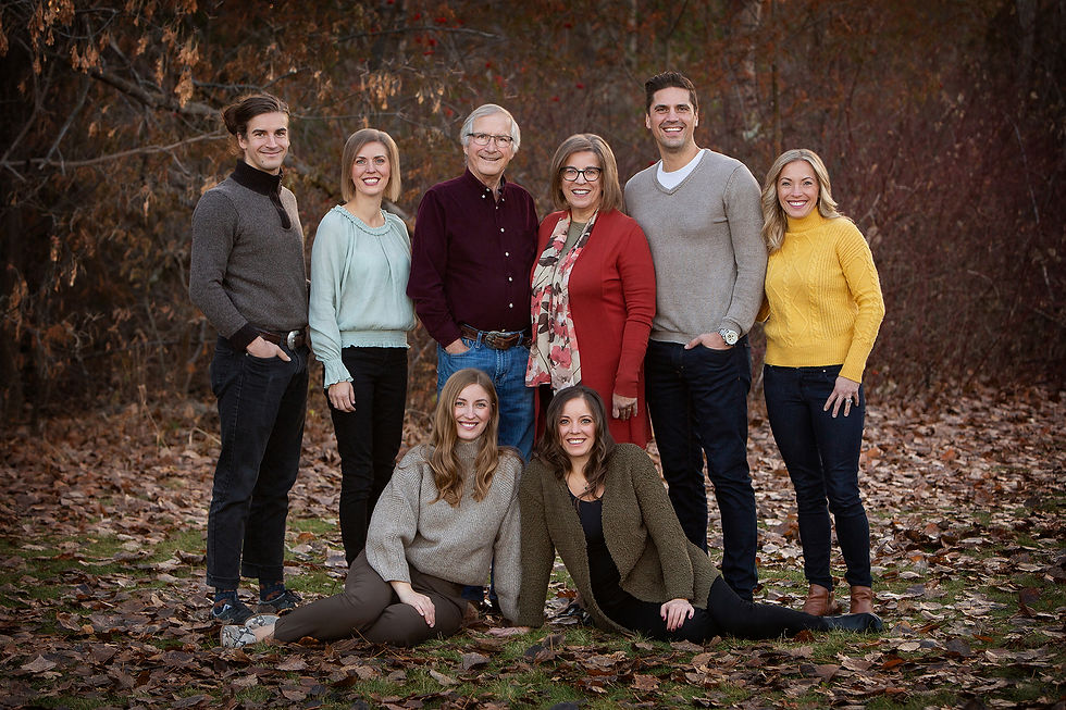 A smiling group of eight stands and sits on fallen leaves, wearing casual fall outfits. The background is wooded, evoking a warm, serene mood. An older couple stands smiling by a wooden fence, surrounded by lush greenery. The man wears a grey shirt; the woman is in a dark sweater. Fall photos in Edmonton.