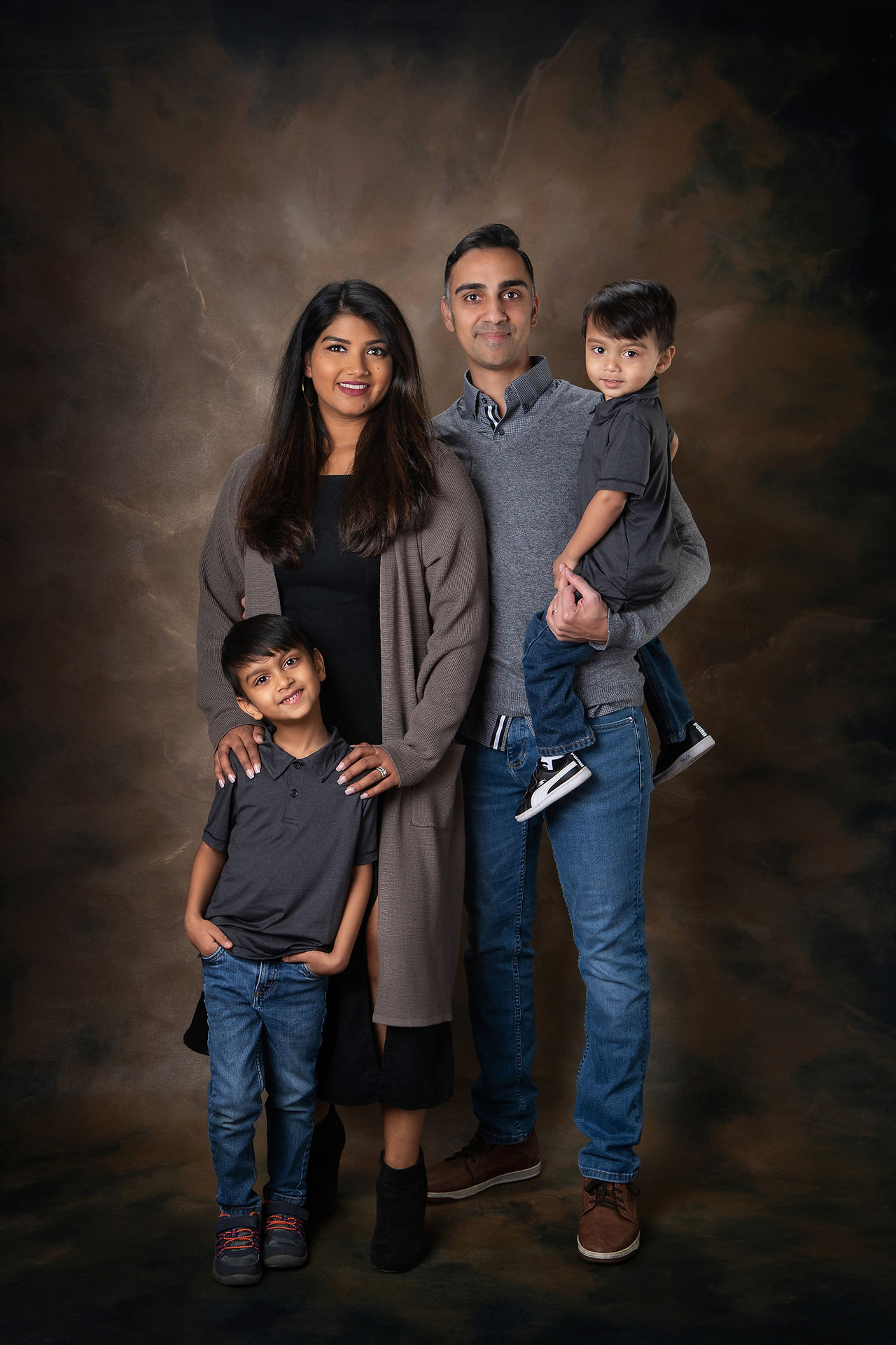 A family of four poses in casual attire. The parents and one child stand while the father holds the youngest child. They are smiling warmly. Edmonton Photographer