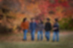 Family of five people walking together, smiling, in a park with vibrant fall foliage in the background. They wear casual sweaters and jeans. Edmonton Photographer