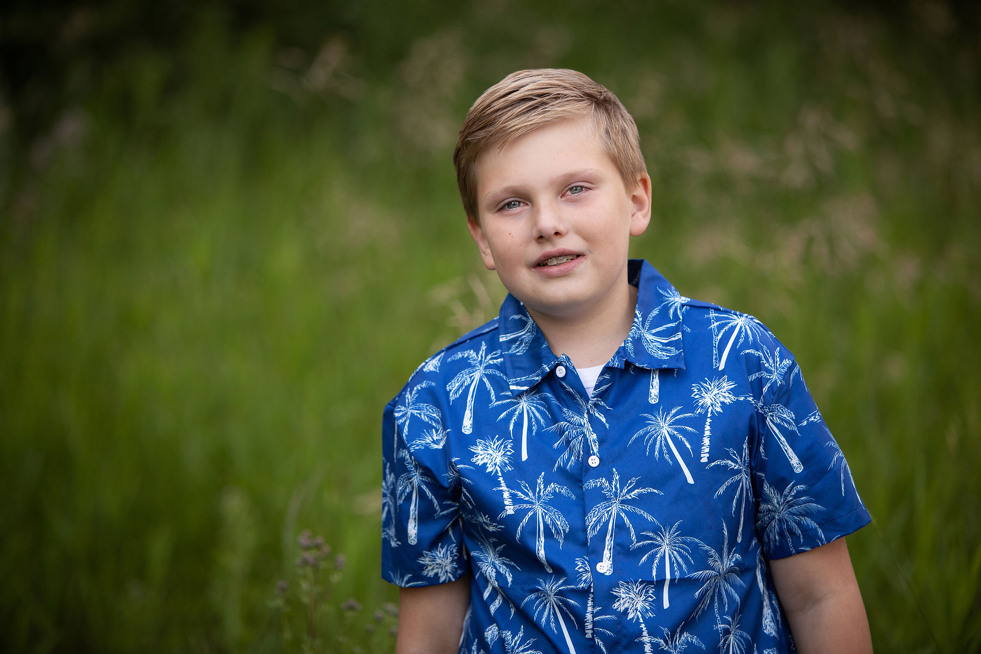 Boy in a blue palm-tree shirt stands outdoors against a blurred green background, looking calmly at the camera. Summer family photos Edmonton
