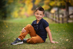 Young boy with short hair in blue polo and orange pants sits on grass, smiling. Blurred autumn trees and a fence are in the background. Children's photography Edmonton