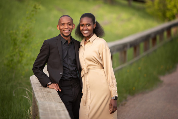 A smiling couple poses outdoors by a wooden railing on a grassy path. The man wears a black suit; the woman is in a beige dress. Viewpoint Photography Edmonton