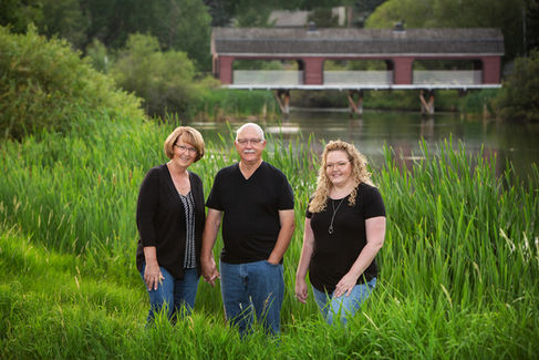 A family of three people stand in tall grass, smiling by a river. A red covered bridge is in the background. They're casually dressed in black tops. Sherwood Park Photographer