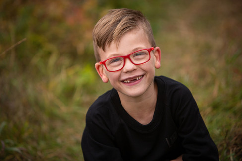 Smiling child with red glasses, wearing a black shirt, outdoors with blurred green background, exudi