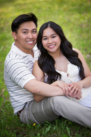 Young couple sitting on grass, smiling. The woman wears a white top, the man a striped shirt. Bright, green background, cheerful mood. Viewpoint Photography Edmonton