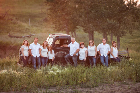 Family in white shirts poses beside a rusty truck in a grassy field at sunset. Trees in the background create a serene, warm setting. Best Edmonton Photographer