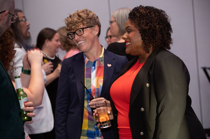 Two people in colourful outfits smile and chat at a social gathering. One wears a rainbow-patterned shirt and glasses. The mood is lively. Viewpoint Photography Edmonton