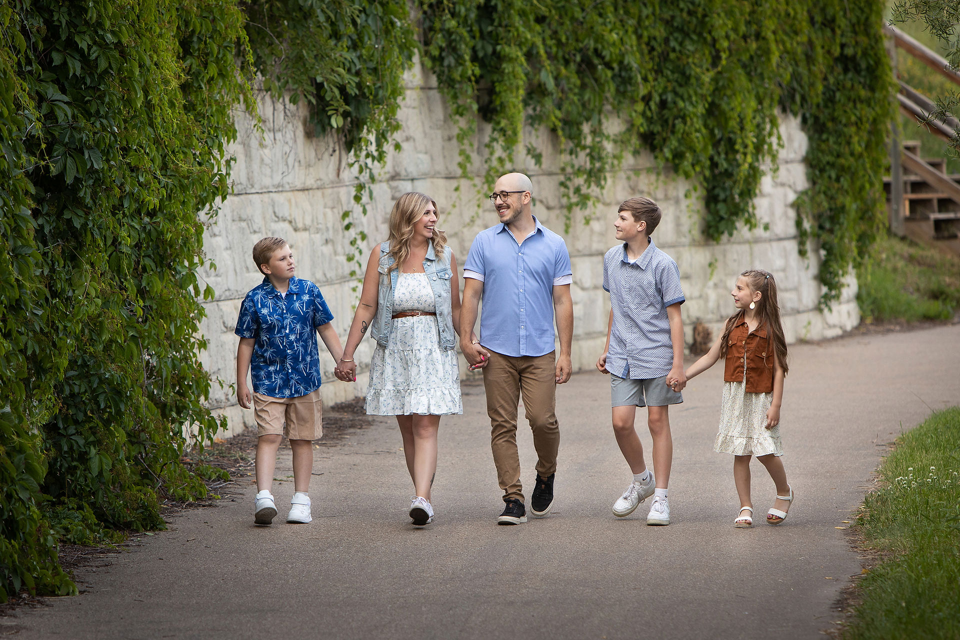 Smiling family of five walks hand in hand on a path beside a vine-covered stone wall in a leafy park. Summer family photos Edmonton.