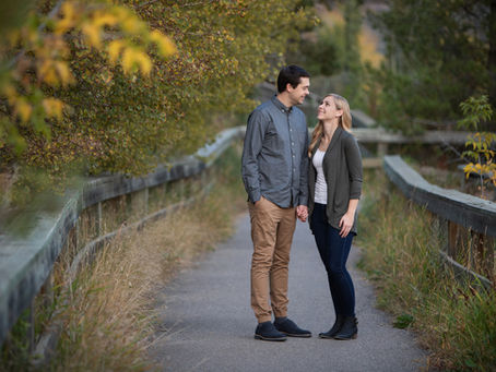 A young couple looking at each other lovingly on a pathway bordered by long grass and a wooden fence.