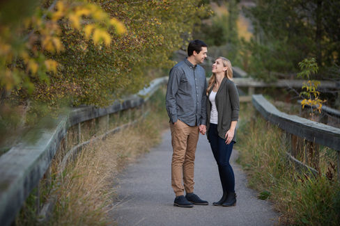 Couple smiling at each other on a tree-lined path, holding hands. Casual attire in earthy tones. Mood is intimate and serene. Edmonton Photographer