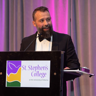 Man in a tuxedo speaks at a podium with "St. Stephen's College" text. Purple-lit curtain backdrop, smiling, formal event ambiance. Viewpoint Photography Edmonton