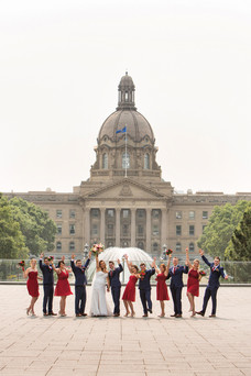 Wedding party in formal attire celebrates with raised arms in front of a grand building with a fountain. Bridesmaids in red, groomsmen in navy. Viewpoint Photography Edmonton