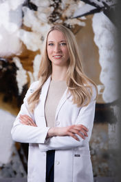Blonde woman in white lab coat with arms crossed, smiling. Abstract brown and white background. Text on coat pocket: "FIGS." Viewpoint Photography Edmonton