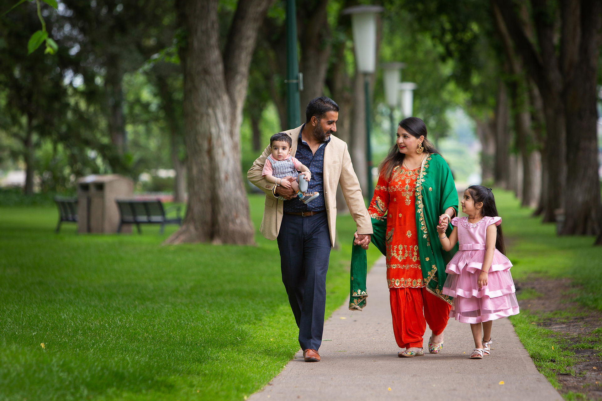 Family of four walking in a park. Man holds baby, woman in traditional attire holds girl's hand. Green grass, trees, and benches in the background.