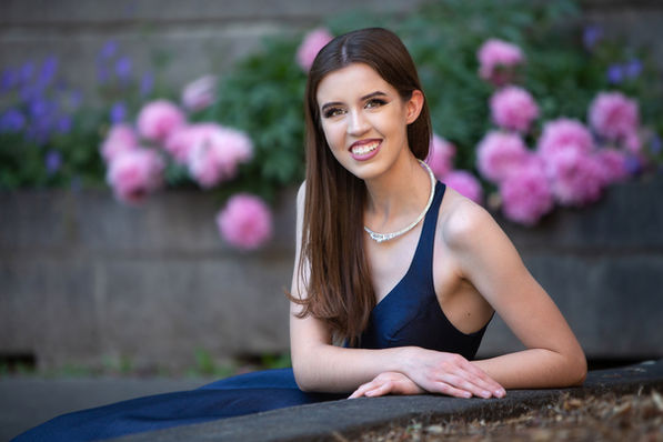 Smiling woman in a dark dress leans on a stone ledge, with pink and purple flowers in the background, wearing a sparkling necklace. Viewpoint Photography Edmonton