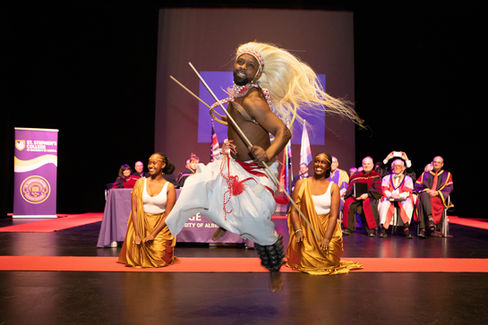 Dancer in vibrant attire leaps on stage, holding sticks. Two seated women in gold dresses smile. Audience and purple St. Stephen's College banner. Viewpoint Photography Edmonton