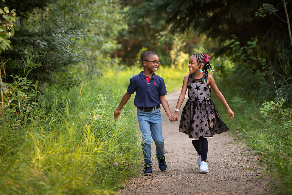 Two smiling children hold hands while walking on a forest path. The girl wears a star-patterned dress, and the boy wears a navy shirt and jeans. Children's photography Edmonton