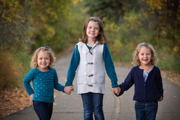 Three young girls smiling, holding hands on a tree-lined path. They're wearing blue tops and jeans, set in a vibrant autumn background. Children's photography Edmonton