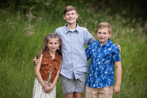 Three smiling children stand together in tall green grass, the older boy with arms around the others, wearing patterned summer clothes. Family photos Edmonton