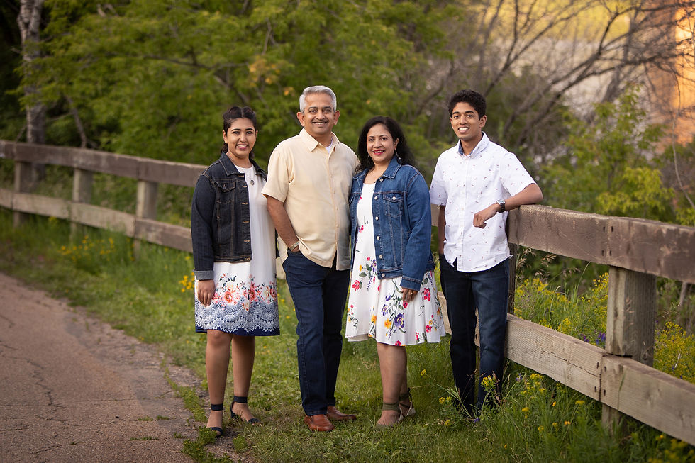 Family of four smiling by a wooden fence, wearing floral dresses and denim jackets. Green trees and sunlit background.