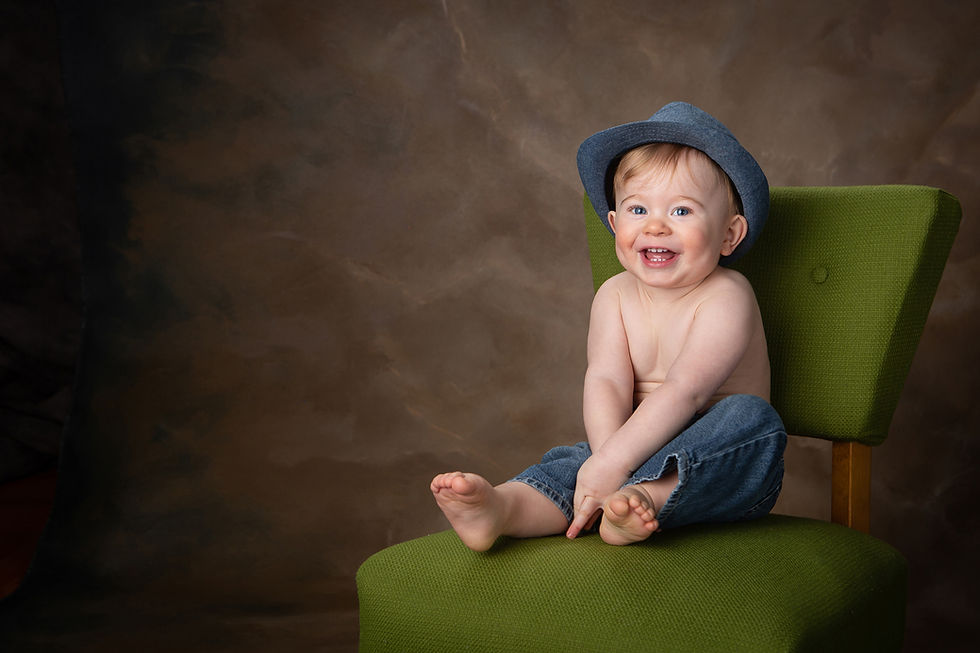 A smiling baby boy sits on a green chir in fron to a brown background. The mood is joyful.