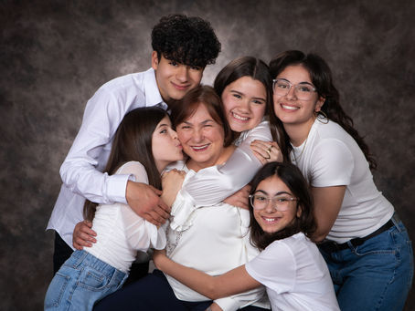 A joyful family hugs a smiling elderly woman. They wear white tops against a dark textured backdrop, conveying warmth and togetherness.