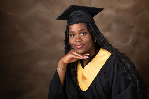 Graduate in cap and gown with yellow stole, smiling against a brown backdrop. The stole displays a logo. Mood is proud and celebratory.