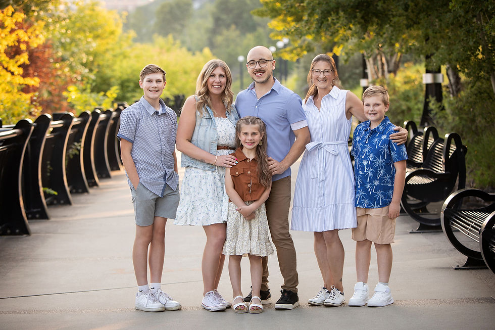 Smiling family of six posing on a tree-lined path with black benches, wearing light summer clothes in warm afternoon light.