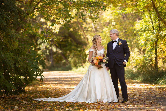 Bride and groom stand on a leafy path, surrounded by greenery. She holds a colourful bouquet, both smiling in a serene, sunlit setting. Viewpoint Photography Edmonton