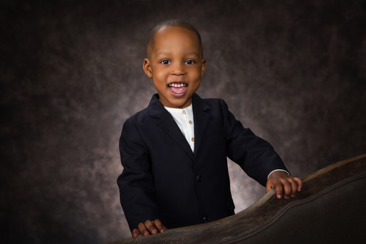 Young child in a navy suit smiles joyfully against a textured dark backdrop, standing with hands on