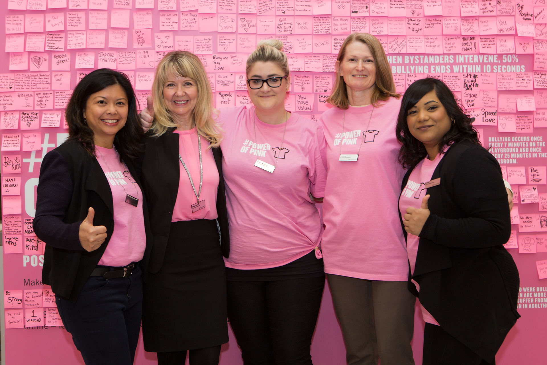 Five people smiling, wearing pink shirts with "Power of Pink" text, stand before a wall of pink sticky notes with anti-bullying messages. Southgate Centre Edmonton Event Photographer.