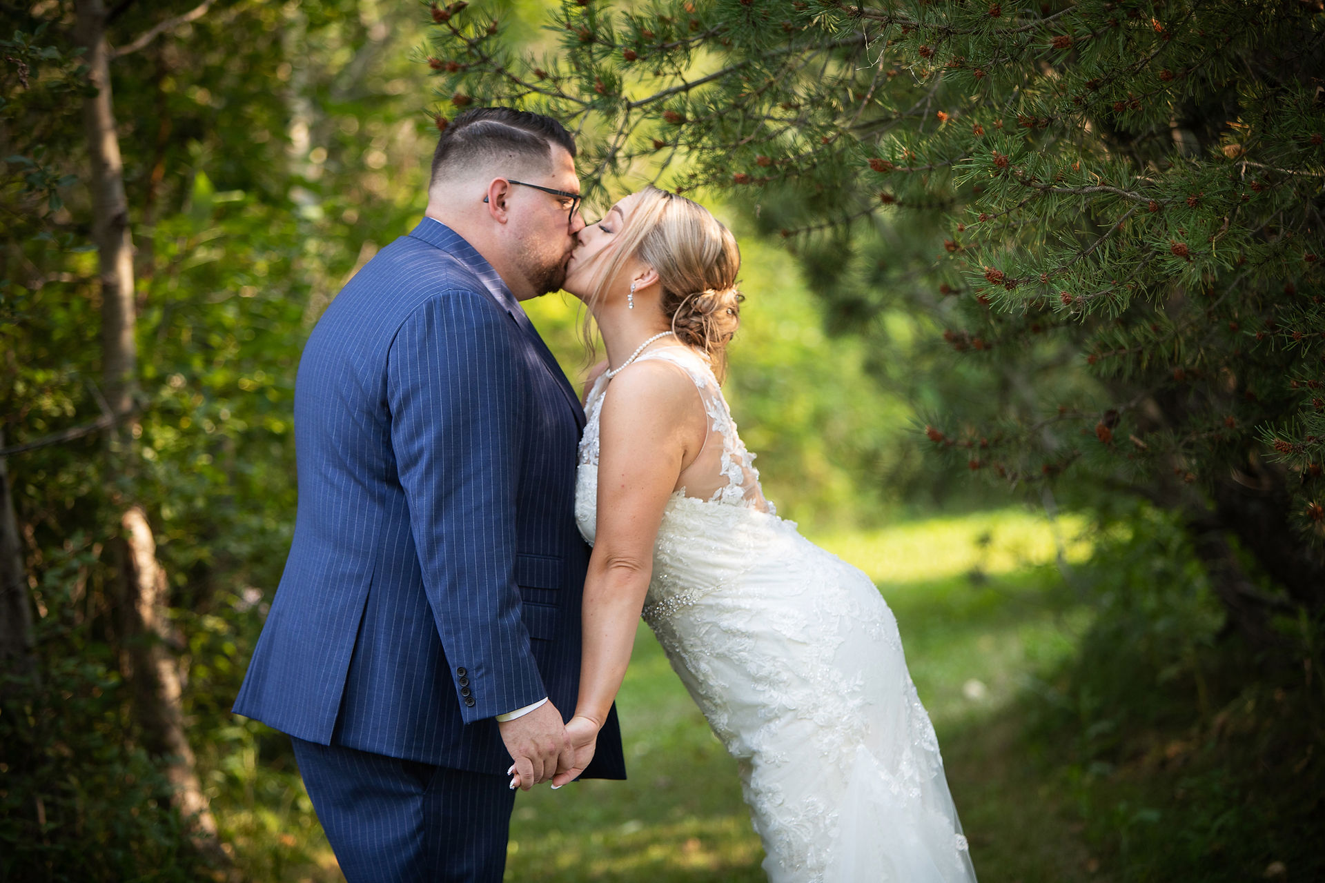 Bride in white dress kisses groom in blue suit on a sunlit forest path, holding hands. edmonton wedding photographer