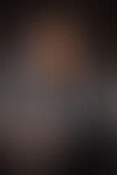 Smiling woman in a grey blazer leans on a dark table against a blurred grey background. Wearing a smartwatch, exuding a professional vibe. Viewpoint Photography Edmonton