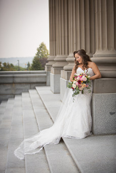 A bride in a white gown holds a colourful bouquet, leaning against stone columns on the wide steps of the Alberta Legislature Building, with trees in the background. Viewpoint Photography Edmonton