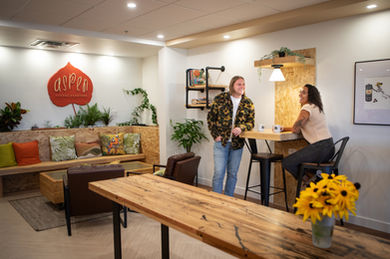Two people chat at a wooden table in a cozy café with plants and colorful cushions. A sign reads "Aspen Coffee Roasters." Mugs on the table. - Edmonton photographer