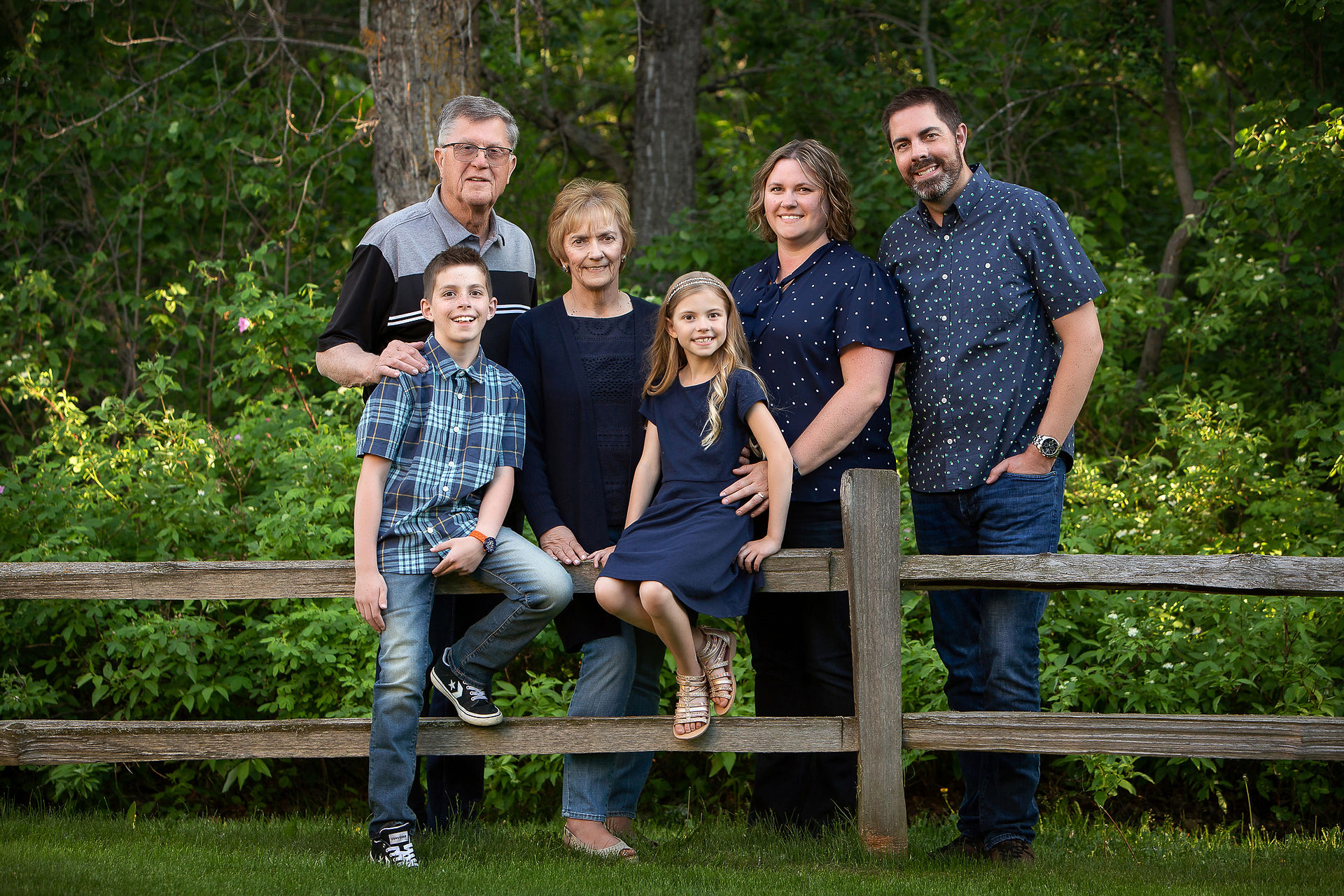 A smiling family of four with two granparents pose by a fence in the park. Family photos Sherwood Park