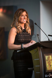 Smiling woman in black dress speaks at podium with Van Gogh art on front. Soft lighting, blurred background creates a formal atmosphere. Viewpoint Photography Edmonton