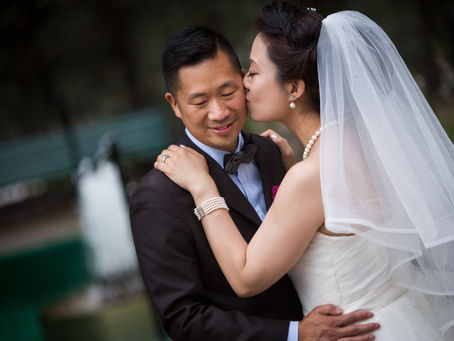 A bride kisses a groom on the cheek as he smiles. A water fountain is in the background. The mood is joyful.