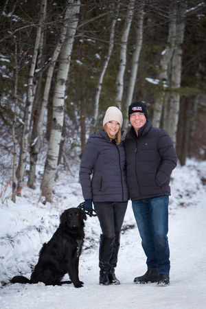 Couple in winter jackets and beanies stands on snowy path with a black dog. Birch trees in background, smiling and joyful mood.