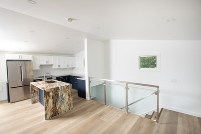 Modern kitchen with granite island, stainless steel fridge, navy and white cabinets, light wood floor, glass railing, and small window. Viewpoint Photography