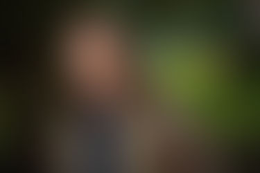 A man with long hair smiles softly, wearing a brown jacket outdoors. The background is blurred green foliage, creating a calm mood. Viewpoint Photography Edmonton