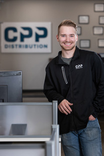 Smiling man in black CPP Distribution jacket, leaning on cubicle in an office. Background shows a CPP Distribution sign. Viewpoint Photography