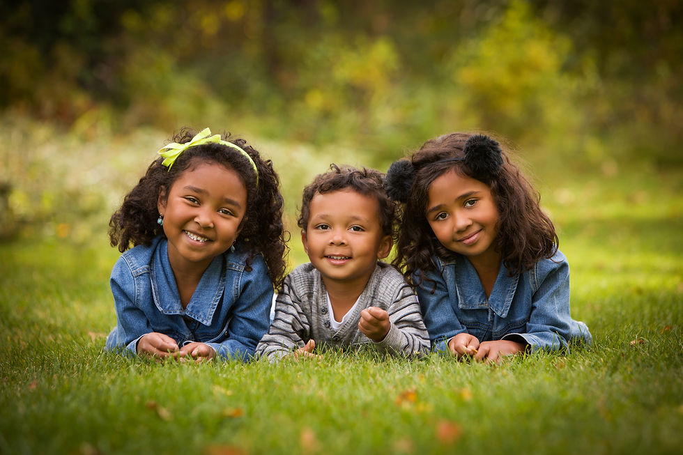 Three smiling children lie on green grass, wearing denim jackets. A girl with a yellow bow, a boy in stripes, and a girl with black earmuffs.