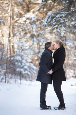 Couple kissing in a snowy forest, both in dark winter coats, holding hands. Snow-covered trees in the background add a serene, romantic mood. Viewpoint Photography Edmonton