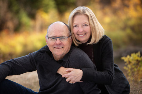 Smiling couple in an embrace outdoors with blurred autumn foliage background. Both wearing dark sweaters, expressing warmth and joy. Fall photo session Edmonton.