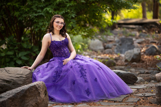Young woman in a purple dress sits on a rock path in a lush garden, smiling. Her gown features intricate lace details and a flowing skirt.